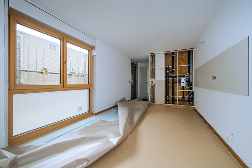 Bild: Wohnraum von innen mit hellbraunem Linoleum, weißen Wänden und weißer Decke und links einem großen, bodentiefen Fenster.