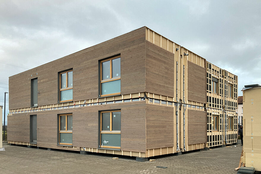 Zwei zweigeschossige Modulbauten auf einem Außengelände, einer davon mit teilmontierter Holzfassade.