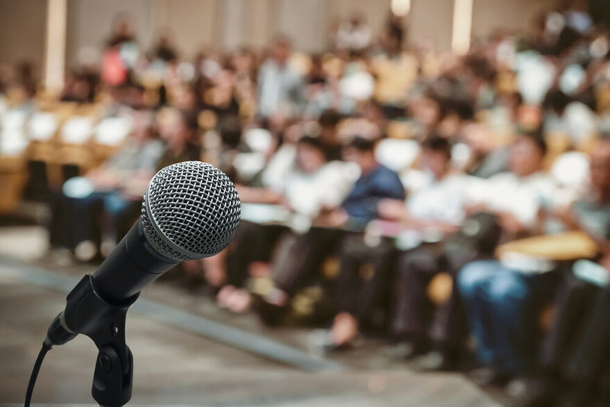 Im Vordergrund ein Mikrofon und dahinter verschwommen viele Menschen, die in einem Saal sitzen