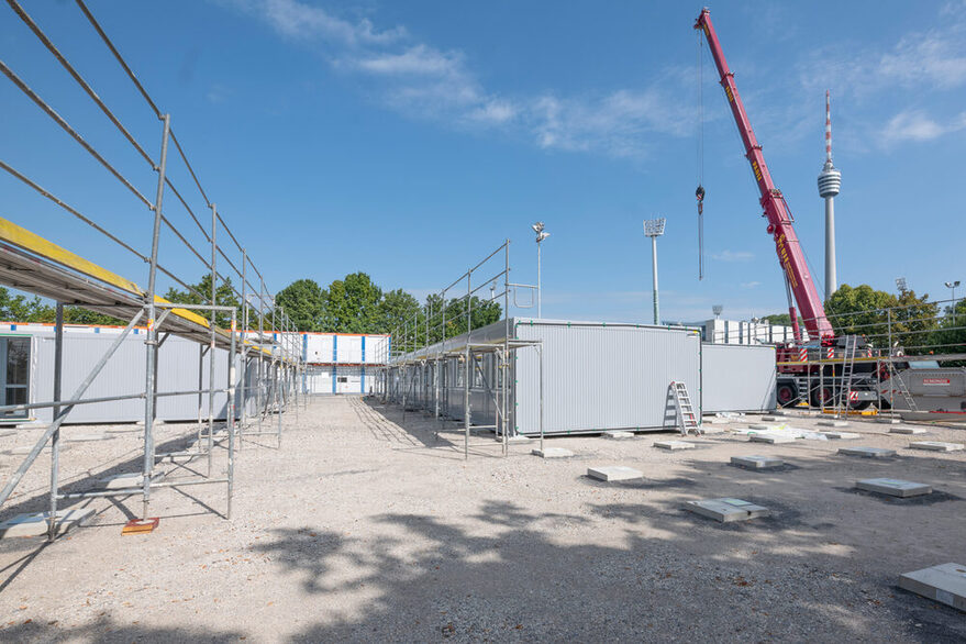 hellgraue, teilweise eingerüstete Container auf einer Kiesfläche und im Hintergrund ein roter Kran