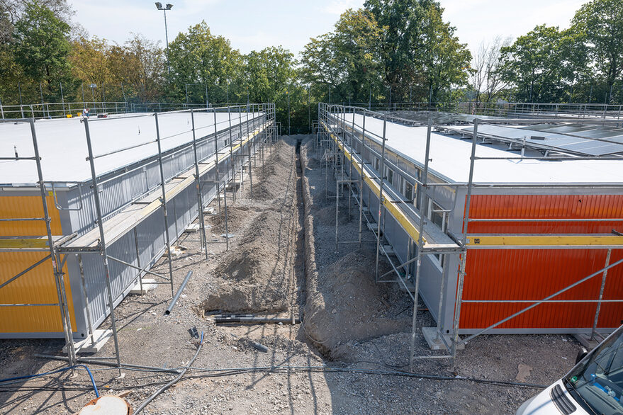 rechts und links jeweils eine Reihe Wohncontainer mit bunten Stirnflächen, dazwischen ein kleiner Graben mit den Rohrleitungen