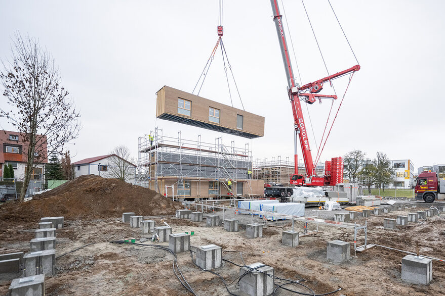 roter Schwerlastkran, der ein mit Holz verkleidetes Wohnmodul vom Lkw an die finale Position hebt