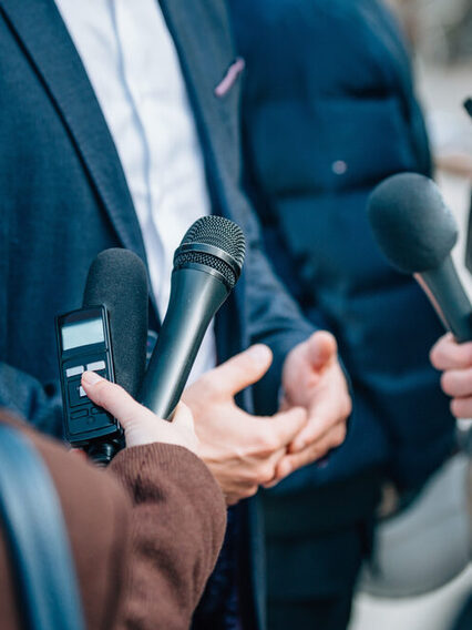Ein Mann im Anzug wird interviewt. Mehrer Mikrofone werden ihm hingehalten.