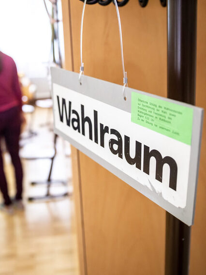 Bürgerinnen und Bürger stehen hintereinander am Eingang vor einem Wahlraum und erhalten ihre Wahlunterlagen.