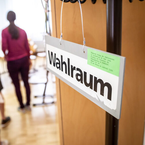 Bürgerinnen und Bürger stehen hintereinander am Eingang vor einem Wahlraum und erhalten ihre Wahlunterlagen.