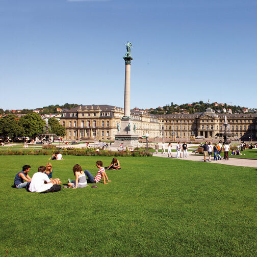 Menschen sitzen auf einer Wiese am Schlossplatz. In der Mitte steht die rund 30 Meter hohe Jubiläumssäule, dahinter ist das Neue Schloss.