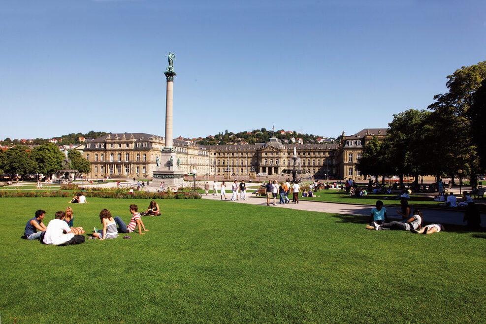 Menschen sitzen auf einer Wiese am Schlossplatz. In der Mitte steht die rund 30 Meter hohe Jubiläumssäule, dahinter ist das Neue Schloss.