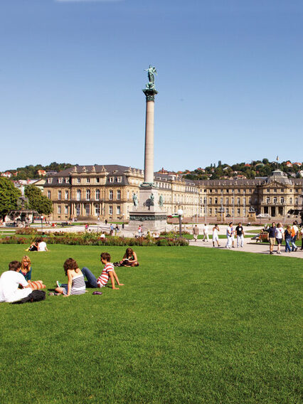 Menschen sitzen auf einer Wiese am Schlossplatz. In der Mitte steht die rund 30 Meter hohe Jubiläumssäule, dahinter ist das Neue Schloss.