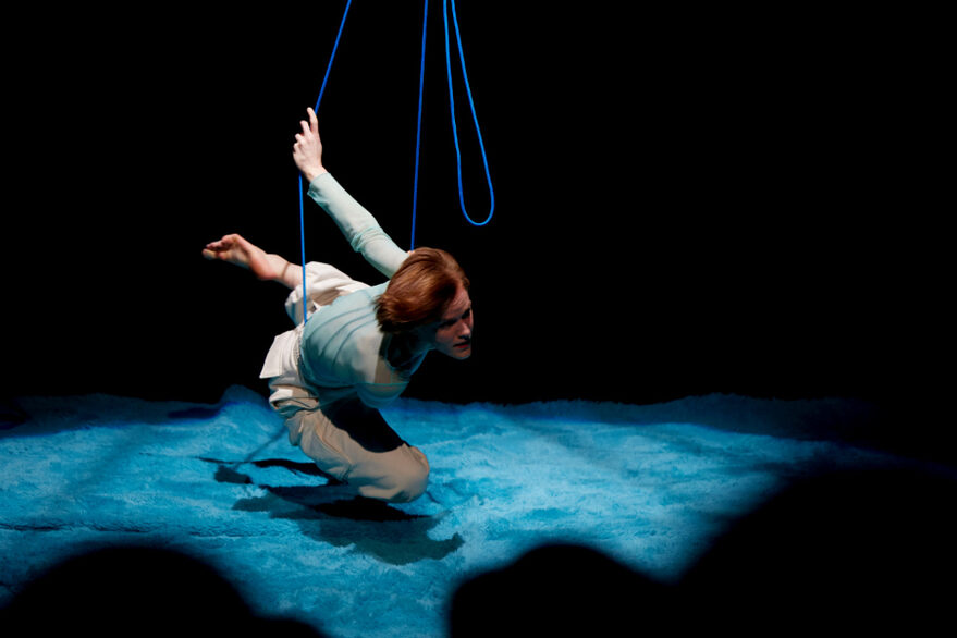 Die Spielerin schwebt, an Seilen aufgehängt über dem blauern Bühnenboden