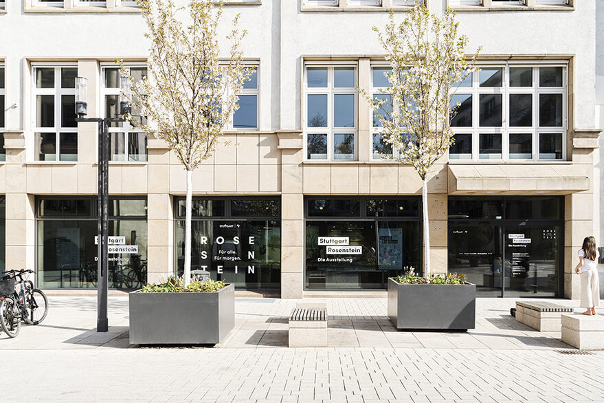 Beige Hausfassade mit vielen Fenstern, im Erdgeschoss beklebt zur Ausstellung. Davor Straße und Bäume.
