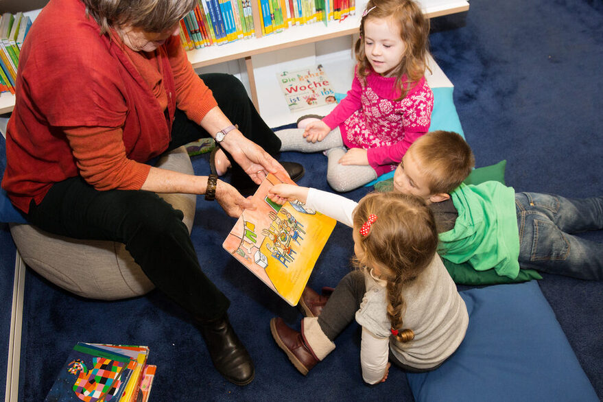 Eine erwachsene Person liest drei Kindern aus einem Buch vor.