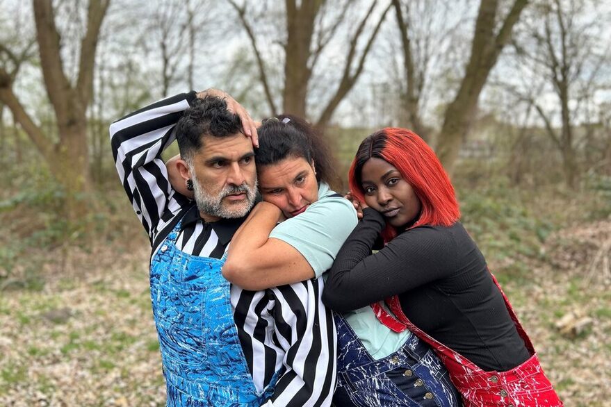 Die drei Künstler*innen der Gruppe "La Fleur" lehnen sich hintereinander aufgereiht aneinander. Sie stehen in einem herbstlichen Wald. Alle drei tragen Latzhosen in verschiedenen Farben. Die Person links hat ihre Hand elegant über den Kopf geschlungen, die anderen beiden haben ihre Köpfe auf ihre Handgelenke gelegt.