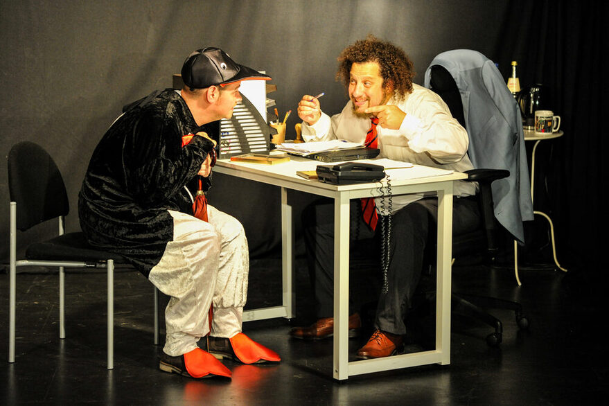 2 Schauspieler an einem Tisch in hitzigem Gespräch