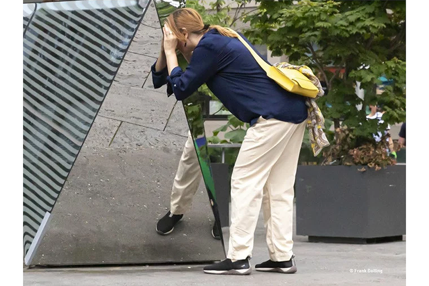Eine Person betrachtet und ertastet ein Kunstwerk mit einem Spiegel. Im hintergrund ist ein Baum in einem Topf zu sehen.