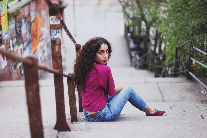 Es ist eine Frau von hinten fotografiert, die auf einer öffentlichen Treppe draußen sitzt und in die sich umdreht, um in die Kamera zu schauen, mit ernstem Blick.