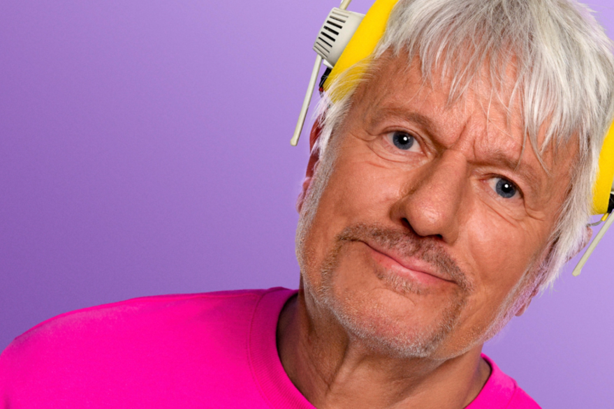 Jürgen Becker în fața unui fundal violet, purtând căști galbene și un tricou roz neon.