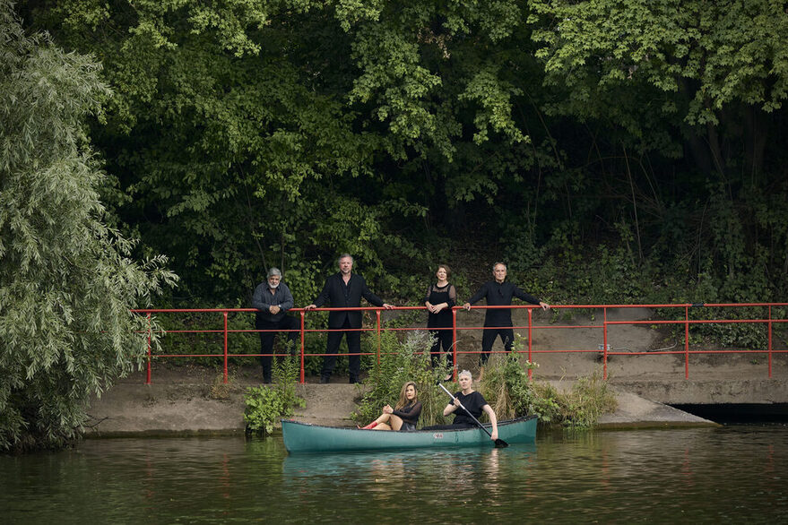 Neue Vocalsolisten. Zwei Personen in einem Kanu und vier Personen am Ufer. (Pressefoto)
