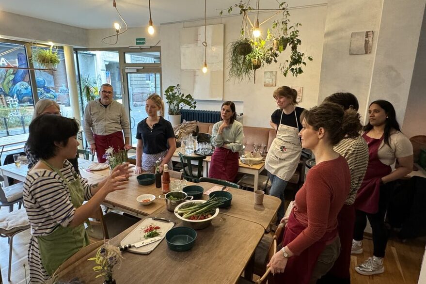 Personen stehen um einen Tisch, hören einer Referentin zu, bevor sie das Gericht gemeinsam kochen.