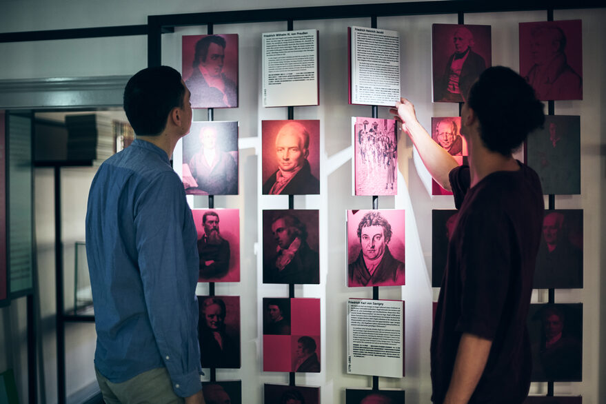 Es ist ein interaktiver Teil der Dauerausstellung zu sehen. Zwei Personen bewegen Kacheln mit Bildern und Texten an einer Wand.