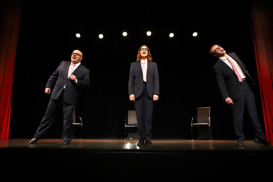 Drei Personen stehen im Anzug auf der Bühne. Zwei Männer und eine Frau. Die Frau steht in der Mitte. Die Männer mit einigem Abstand links und rechts von ihr.
