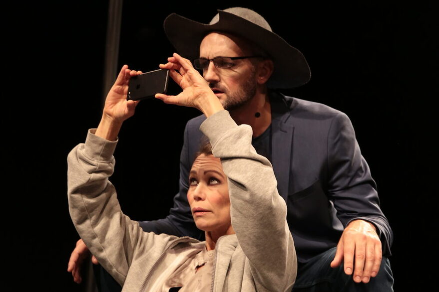 Szenenfoto - Zwei Personen auf der Bühne, eine sitzt und hält einen kleinen schwarzen Gegenstand in die Luft, die andere Person steht dahinter.