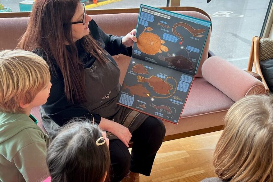 Kinder hören einer Frau beim Vorlesen aus einem Bilderbuch zu.