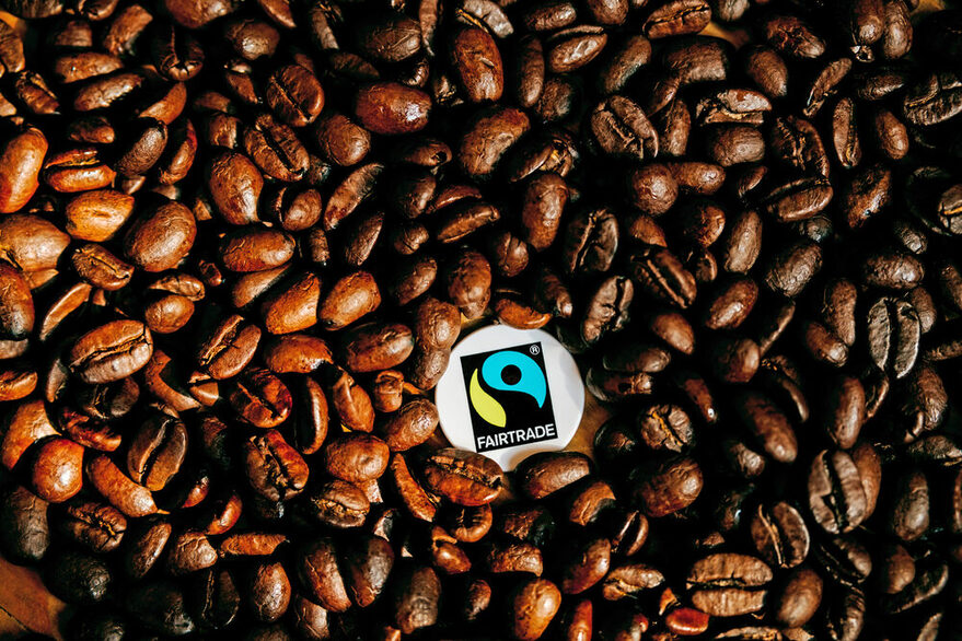 Weiße Brosche mit Faritrade-Siegel liegt auf schwarzen Kaffeebohnen.