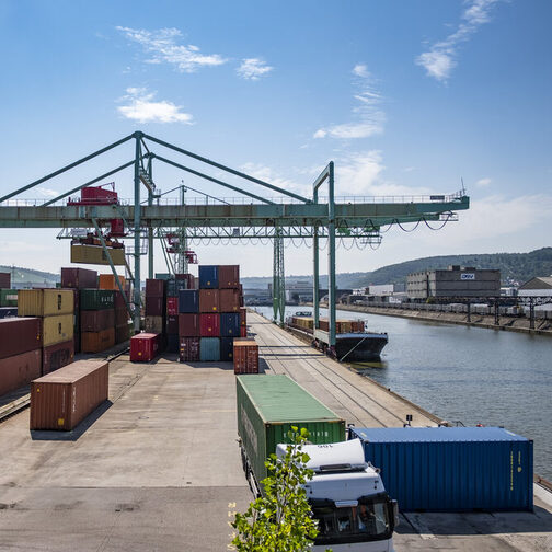 Blick auf ein Hafengelände am Flussufer mit vielen bunten Containern und einem großen Kran.